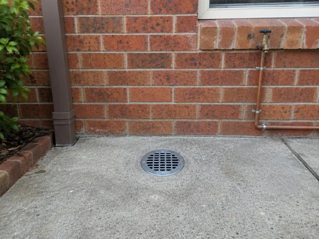 Outdoor drain grate set in dry concrete paving beside a brick wall and copper pipe in Miranda